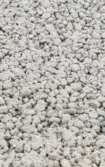 Nahaufnahme einer Oberfläche aus Drainagebeton. Die Struktur besteht aus vielen groben, rundlichen Gesteinskörnern, die von einer dünnen Schicht Zementleim umhüllt sind. Zwischen den Steinen sind deutlich sichtbare Hohlräume, wodurch der Beton offenporig und wasserdurchlässig wirkt. Die Textur ist unregelmäßig und rau.

                                    